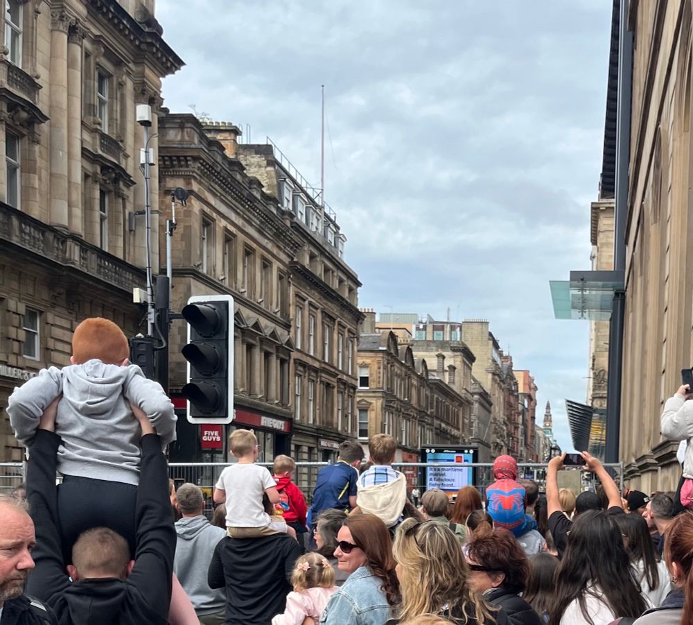 Crowd of people with children on parents’ shoulders, including one child in a Spider-man costume, ahead of barriers on a Glasgow street 