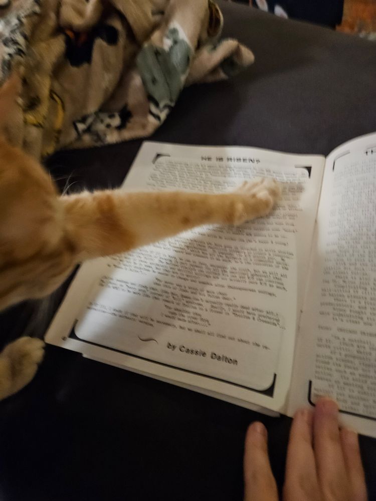 Photo of my orange cat where he's a little blurry from movement because he's reaching out to stretch his paw on my fanzine. Or hold the page down for me while I try to take photos? He's so helpful.