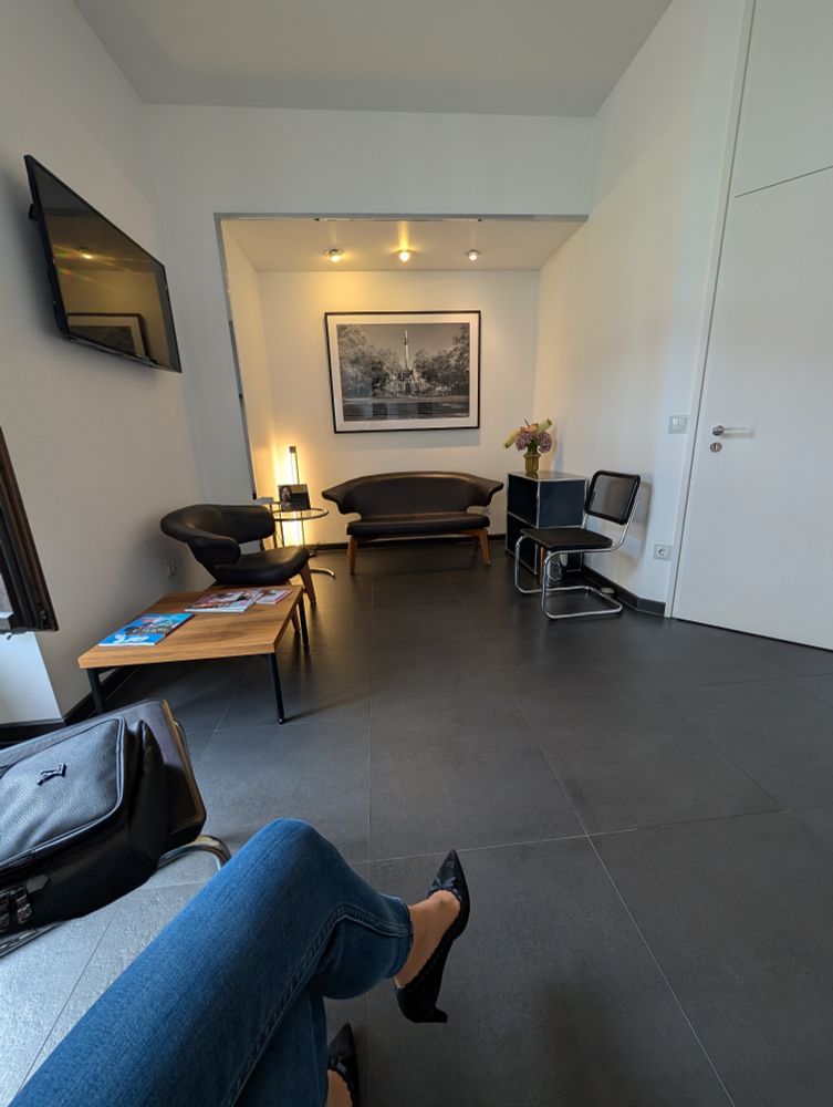 Ein Wartezimmer mit einem dunkelgrauen Fliesenboden. Eine Ledersitzgarnitur und kleine Tische. Die Wände sind weiß. Links hängt ein kleiner Fernseher. An der anderen Wand ein großes eingerahmtes S/w Foto. 

Ach ja und die Beine der Userin in blauer enger Jeans. Sie trägt schwarze Pumps. 