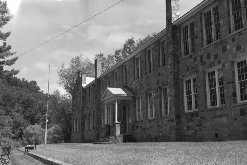An monochrome photograph of a wide, monolithic two-storey stone building with many windows and a single, narrow doorway set amid manicured lawn and parkland.