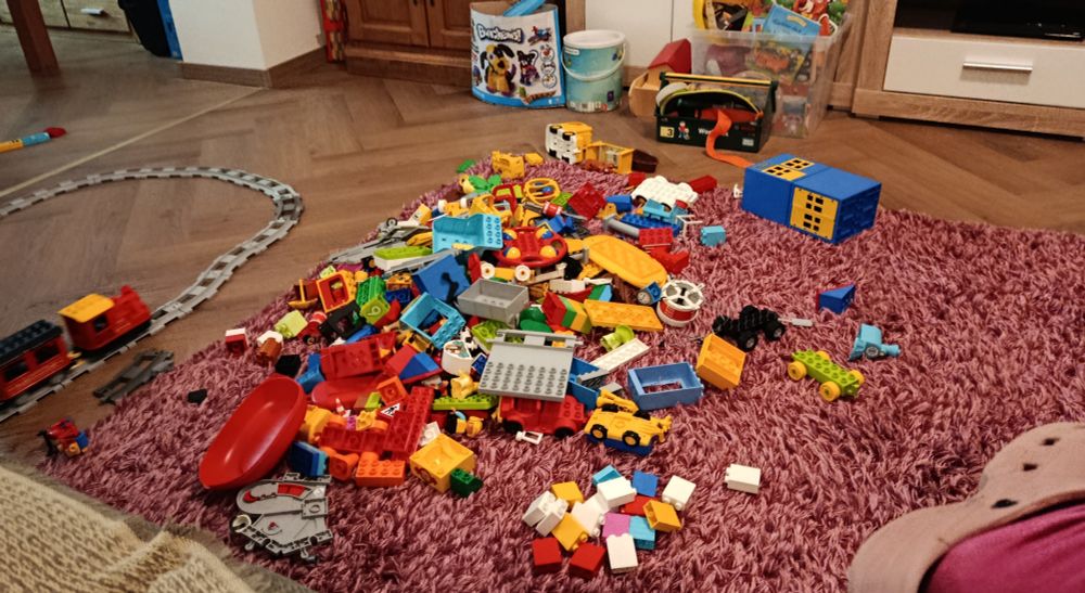 Der Fußboden ist voller bunter Lego Duplo Steine. Am Bildrand links fährt eine kleine, rote Eisenbahn auf grauen Schienen.
