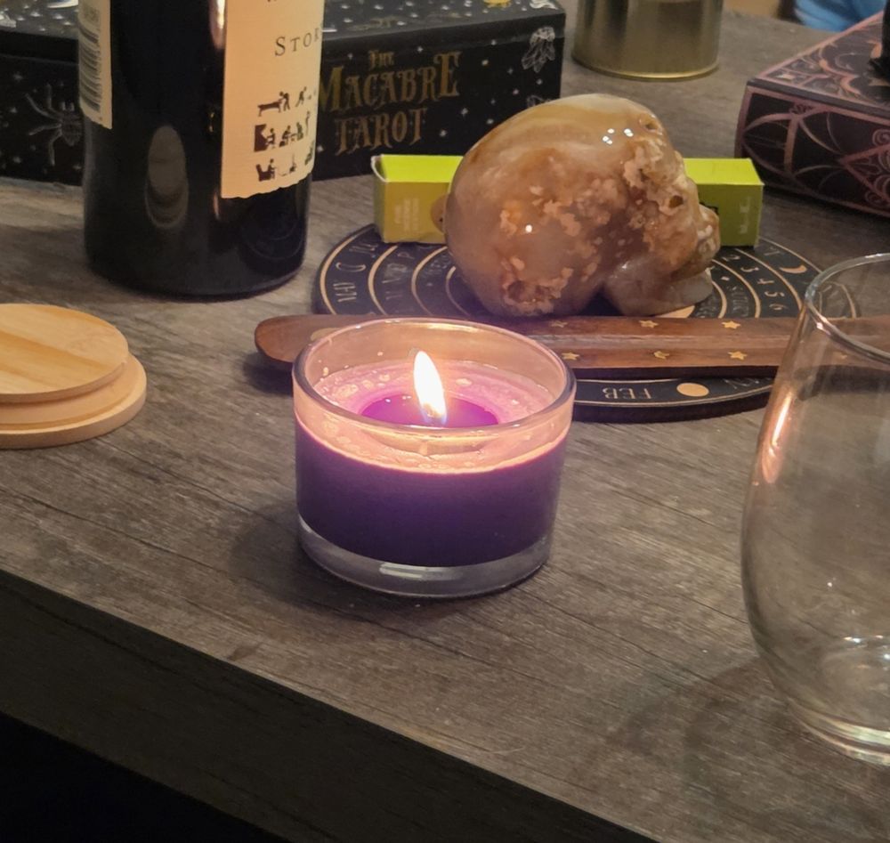 A short burning jar candle surrounded by a bamboo lid, a wine bottle, a flower agate skull on a pendulum board, a wine glass, a macabre tarot deck, and a few fake books. 