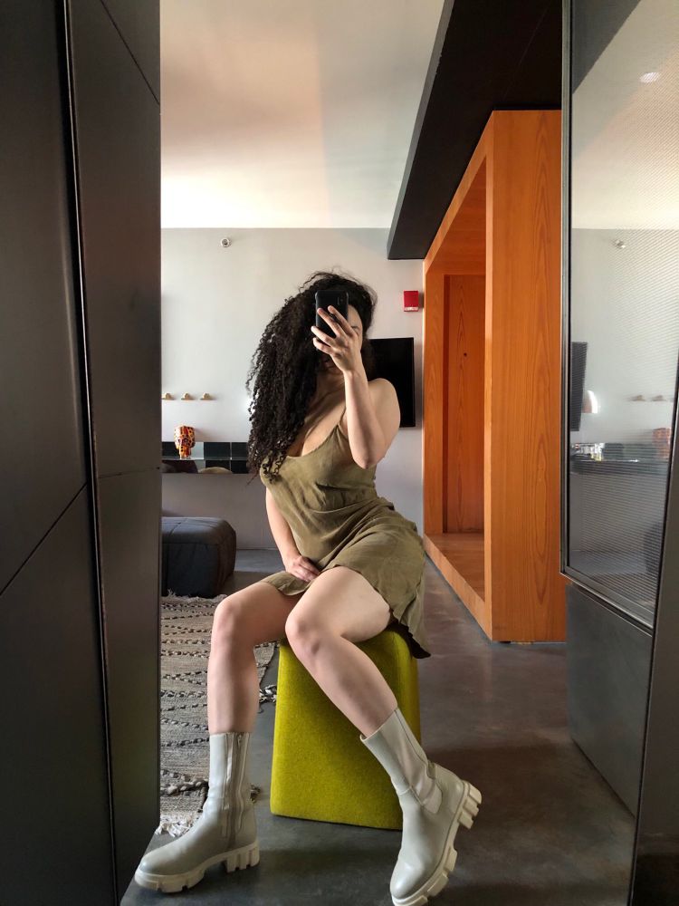 A mirror selfie of Lila, light skinned sensuously curvy and toned, wearing a short olive slip dress and chunky grey leather boots sitting with legs slightly parted on a chartreuse felted stool with long voluminous brown curls cascading over one shoulder sitting in a room of clean lines. Soft light, warm wood, moody concrete, sleek metal hand woven textile.