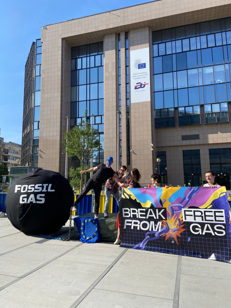 A group of people in front of a colourful banner that says Break Free from Fossil Gas pulling a person dressed as the EU chained to a big prisoner’s ball that says fossil gas 