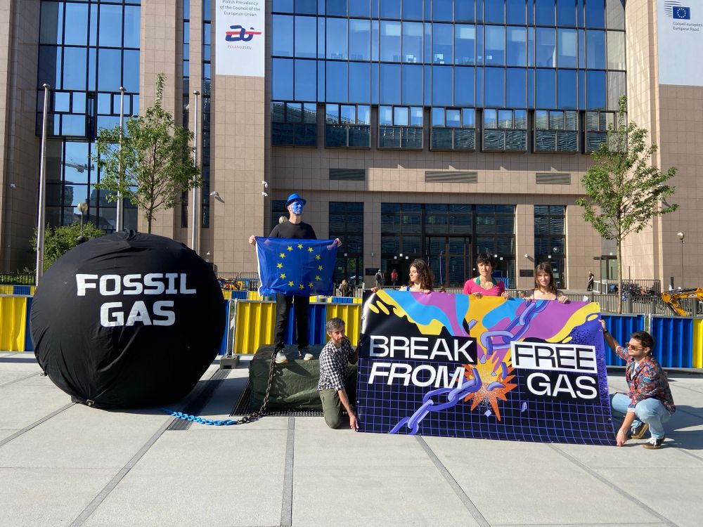 A group of people in front of a colourful banner that says Break Free from Fossil Gas next to a person dressed as the EU chained to a big prisoner’s ball that says fossil gas 
