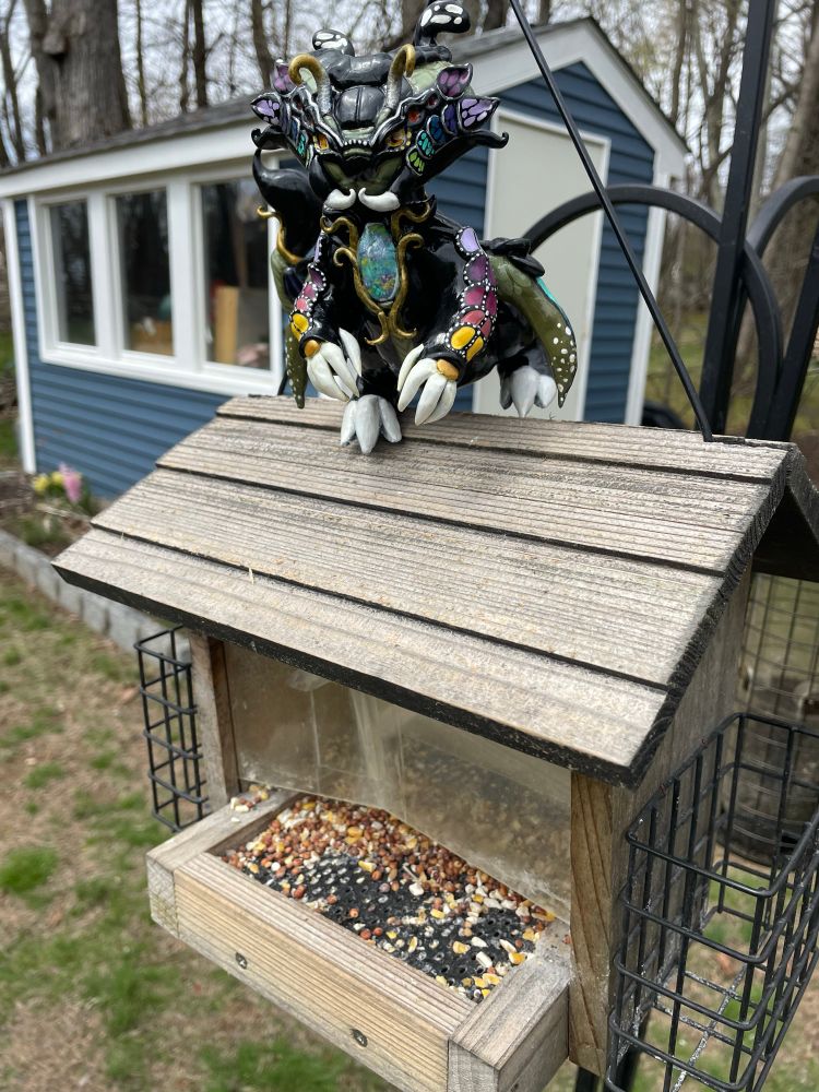 A photo of Peeko, a sculpture of a dragon-like creature, sitting on top of a bird feeder. Behind the bird feeder are trees and a shed with blue siding. 