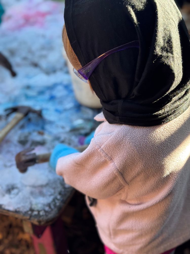 Child in a pink sweater, a black balaclava, and wearing plastic protective glasses holds a hammer at a table covered in ice outside.