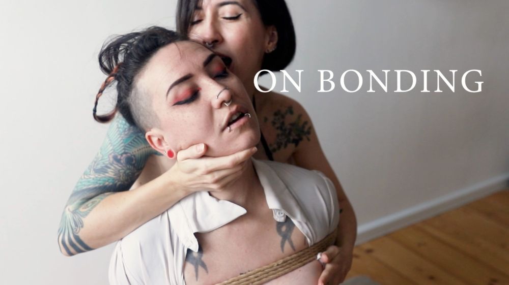 Poster for the short movie : On Bonding
A serene white person with a white shirt and braids is tied up and held by a brunette smiling.