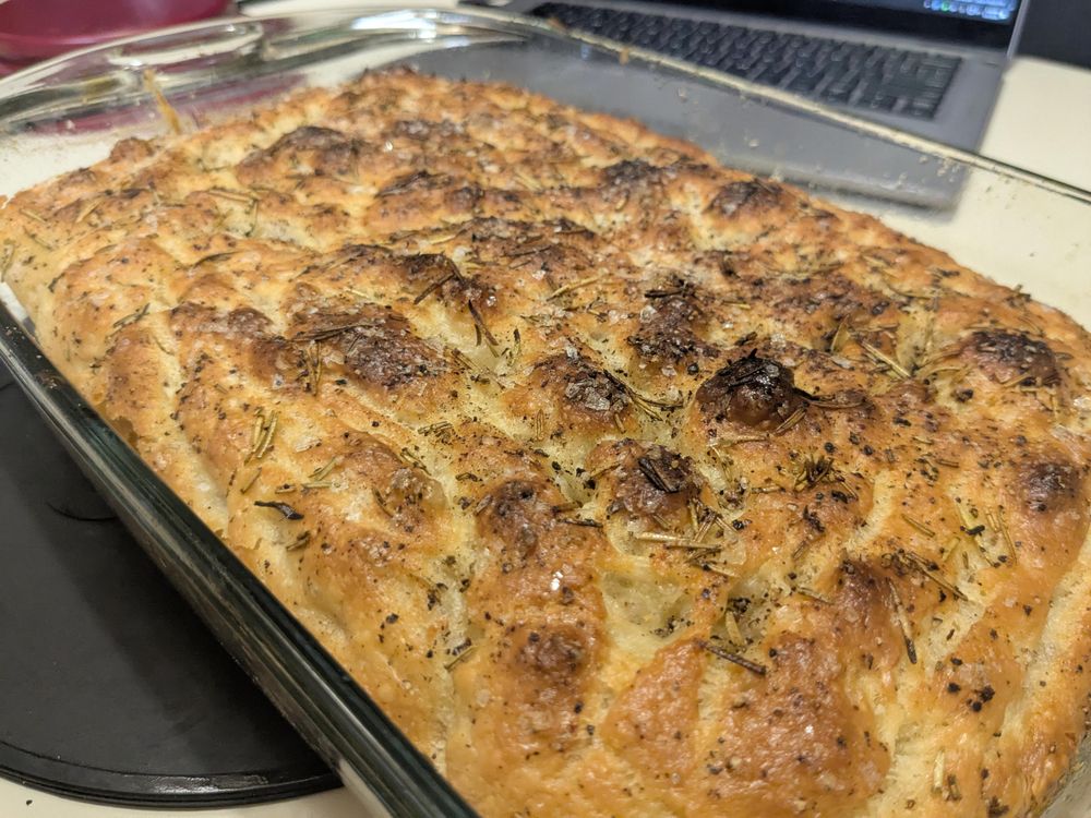 Photo at an angle over a 9-in x 13-in pan of focaccia bread. It's beautifully blistered on top with that signature "fried" look of freshly baked focaccia loaves, scattered with coarse toppings well. There's a laptop in the background because of streaming.