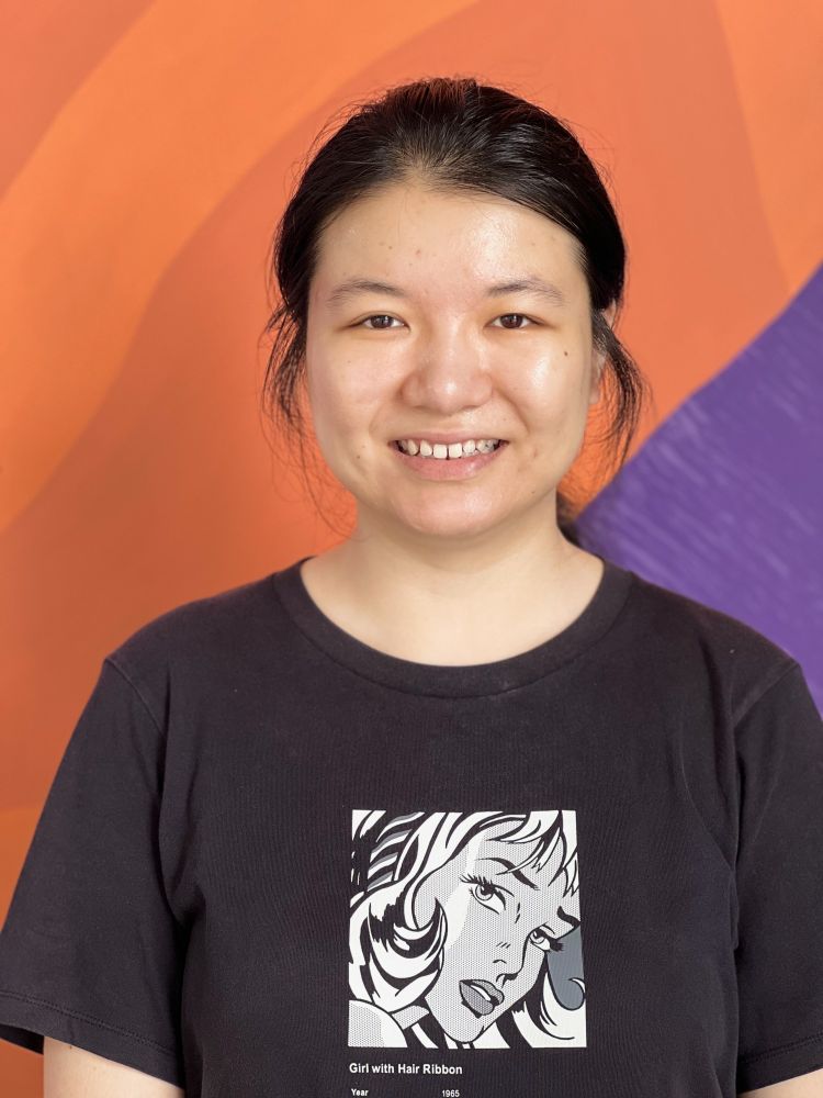 A photo of a young woman smiling directly to camera. She's standing in front of a brightly painted wall in shades of orange and purple. She has dark hair that's been tied back, with a few strands loose over her ears. She's wearing a black t-shirt with a grey and white pop-art graphic of Girl with a Hair Ribbon.