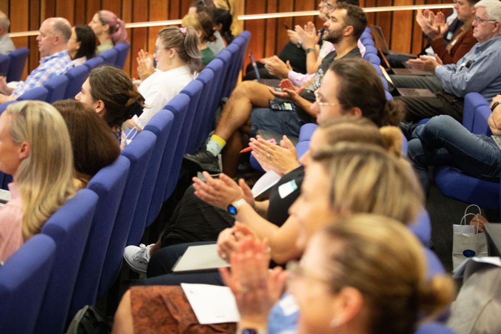 An image featuring a large group of people in a lecture theatre smiling and applauding. The seats are a purplish-blue, and the walls are vertical timber. There is a broad mix of people in the room