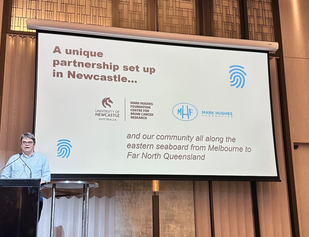 A man stands at a lectern before a large screen with a slide on it. The slide reads, a unique partnership set up in Newcastle... with two logos - the MHF Centre and the MHF. Followed by the words and out community all along the eastern seaboard from Melbourne to Far North Queensland.