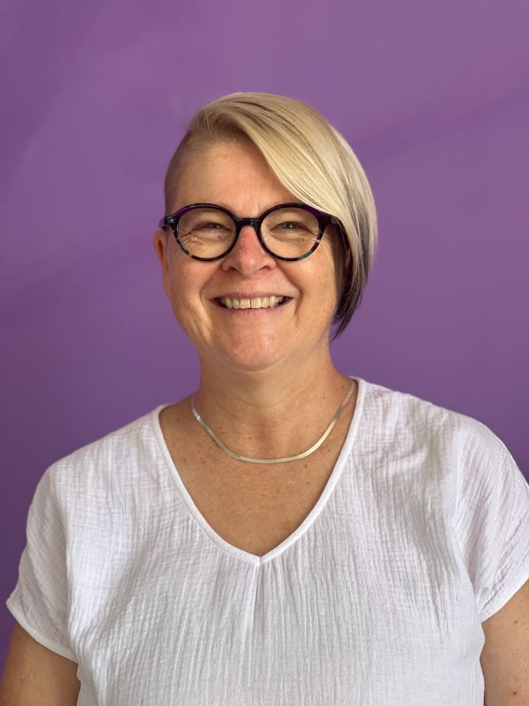 An image of a woman smiling directly to camera. She has styled blonde hair, with streaks of black, and buzz cut on one-side. She wears glasses and a textured white top with a v neck and a silver necklace.