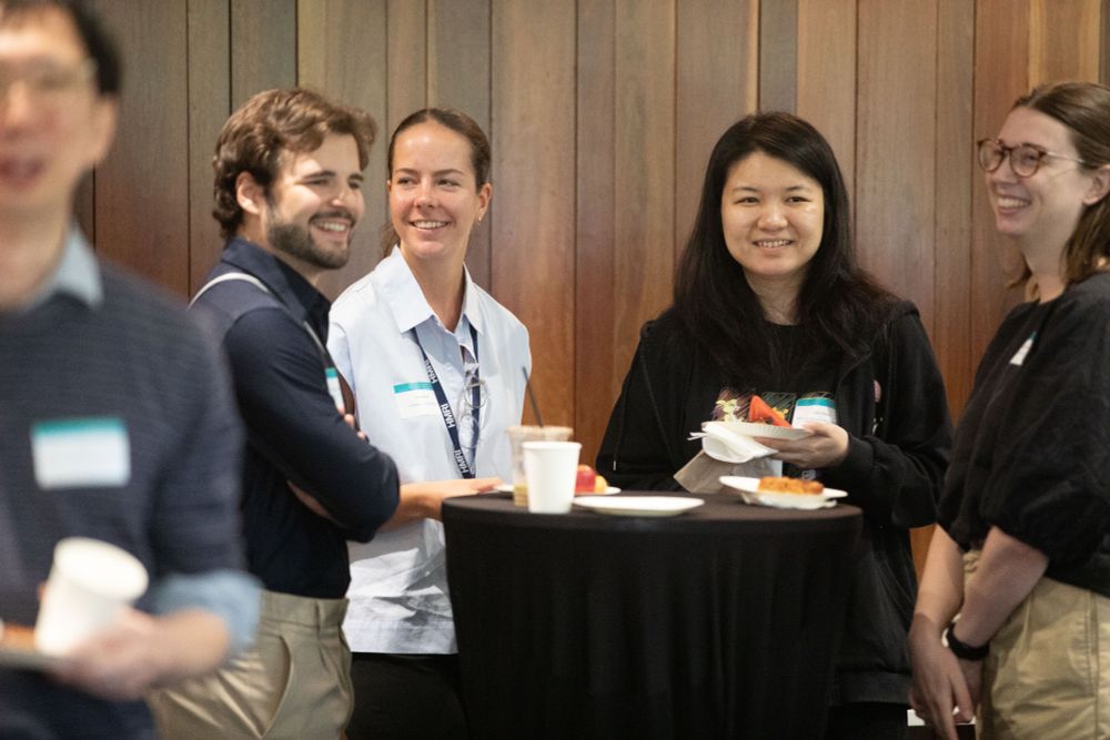 An image featuring five people, all smiling and engaged in conversations in the room. From left: a man is walking to camera. He wears glasses, a collared shirt and blue-grey jumper. Next, a young, bearded man is smiling and engaged, looking off to the right of the photo. Next, a young woman wearing a pale, collared shirt. Her hair is mid-brown and tied back. She is smiling and looking to her left. Next a woman with long, below shoulder-length dark hair hair is smiling face-on to camera. She is wearing a dark top and stands behind a table. Far right: a woman is smiling/laughing in profile. Her hair is deep chestnut and she is wearing glasses and a black top/khakki pants. 
