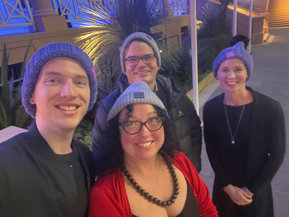 Four people are pictured outside NSW State Parliament which is lit up in purple and gold. They are all wearing MHF Beanies for Brain Cancer. From left, man in black is smiling at the camera and wears a lavender beanie. Next, woman with red cardigan and beads and glasses is wearing a grey beanie. Behind her, a man is smiling and wearing a grey beanie and a puffer jacket. Right a woman dressed in black smiles at the camera. She is wearing a lilac beanie.