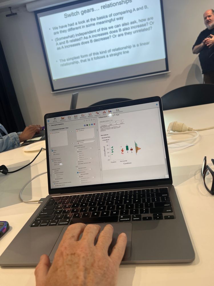 In the foreground is a laptop with text on the screen demonstrating statistics. A hand is on the touchpad, and a pair of black framed glasses are off to the righthand side. On the left, you can see another pair of hands working a laptop. In the background (but the main part of the pic) you see a screen with text explaining statistics. Off to the side is a bearded man with a black t-shirt who is presenting to the room.
