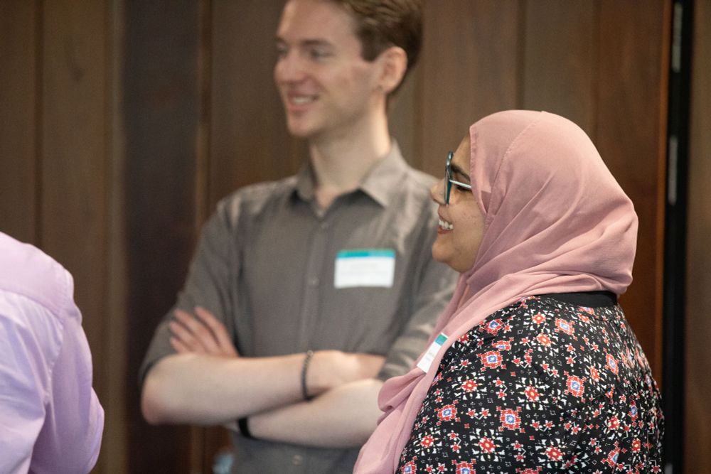Two people are featured in conversation. At the rear of the photo a young man stands, smiling at someone off camera. He's wearing a grey/green collared shirt, rolled to the elbows and has his arms gently folded. In the foreground is a young woman, smiling in profile. She is wearing a rose-coloured headscarf and dark background floral top.