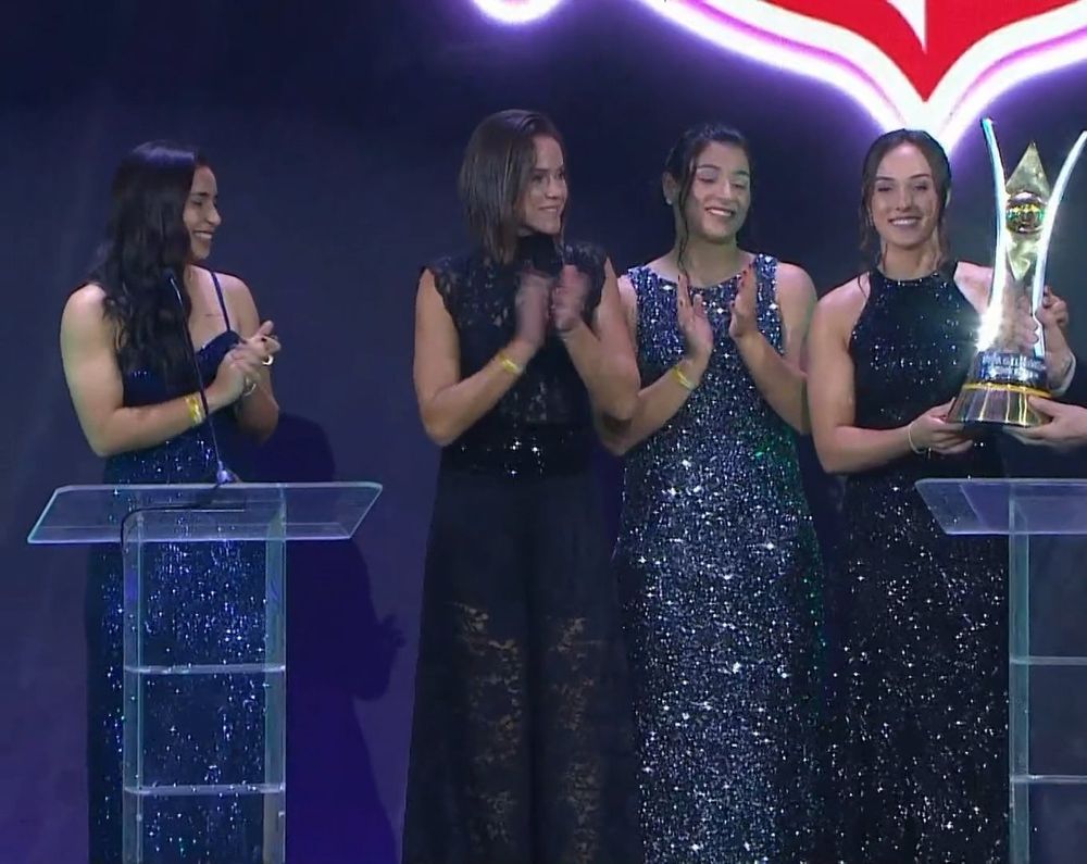 Jogadoras Mariza, Erika, Duda Sampaio e Nicole do Corinthians no palco do Prêmio Brasileirão 2025. Todas estão com vestidos longos de festa. Nicole segura um troféu.