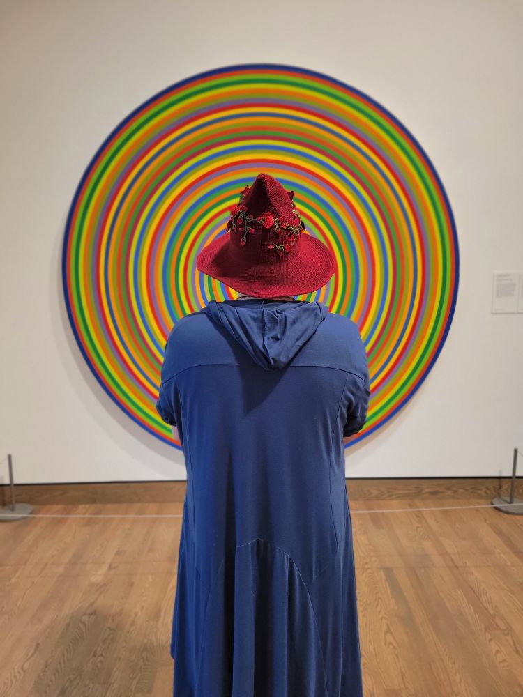 A figure in a blue light robe stands in front of a circular painting in a red hat. 