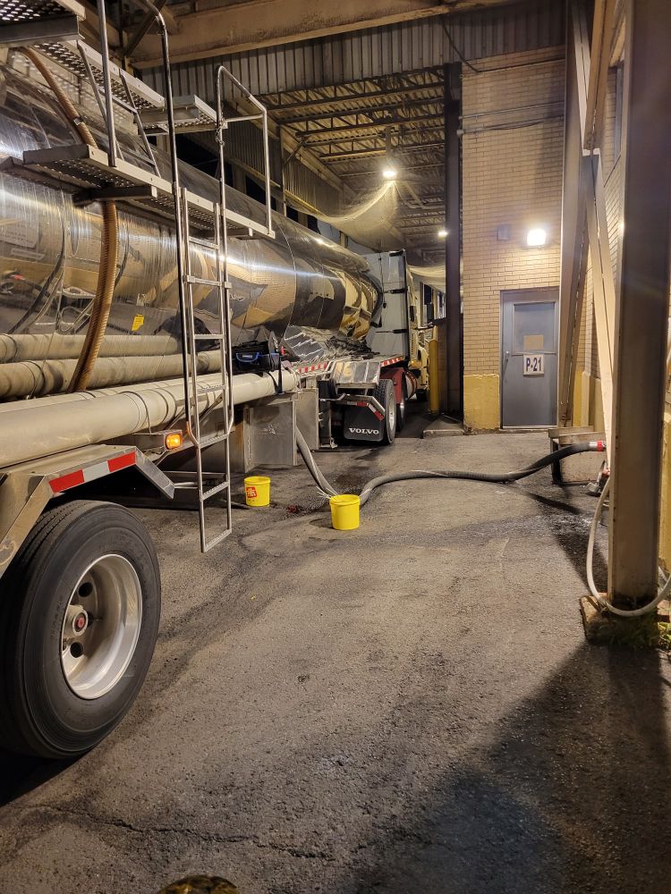 A large silver tanker is connected to a building via a hose. There are yellow buckets on the ground. 