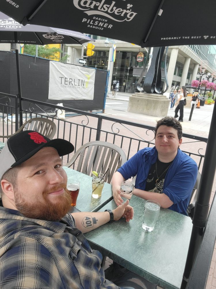 Two gays sit at a patio table in a city. 