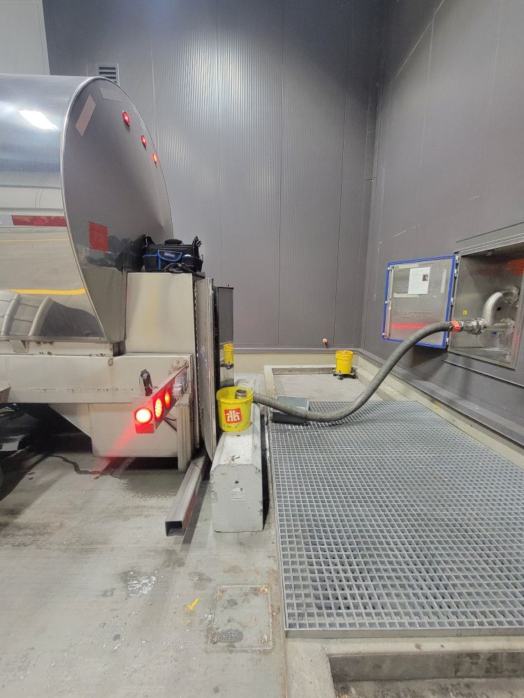 The back of a tanker trailer hooked up to some plumbing on a wall with a hose, with a yellow bucket next to it. 