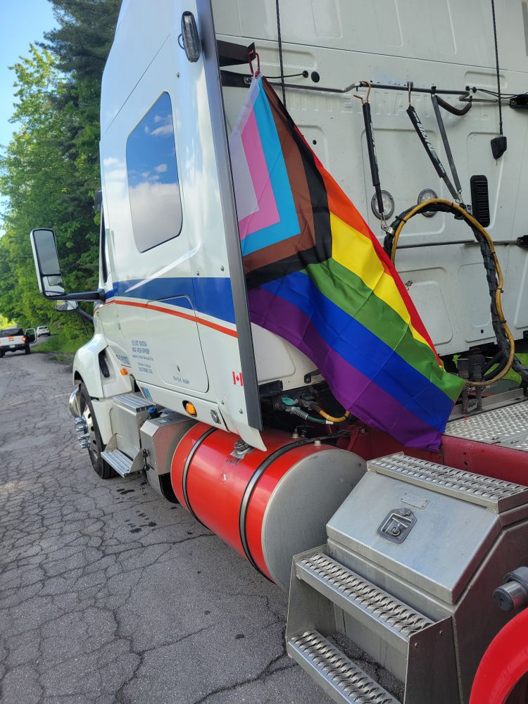 A white semi truck with a inclusive pride flag mounted to the fairing behind the cab.
