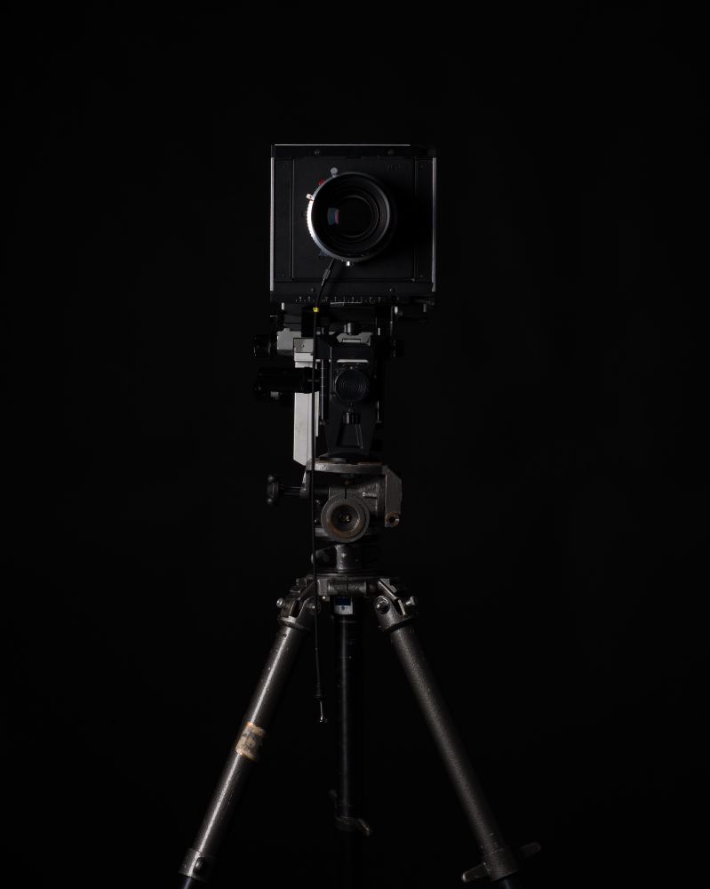 a large format monorail camera on a heavy tripod, lit with studio lights against a black colorama backdrop.