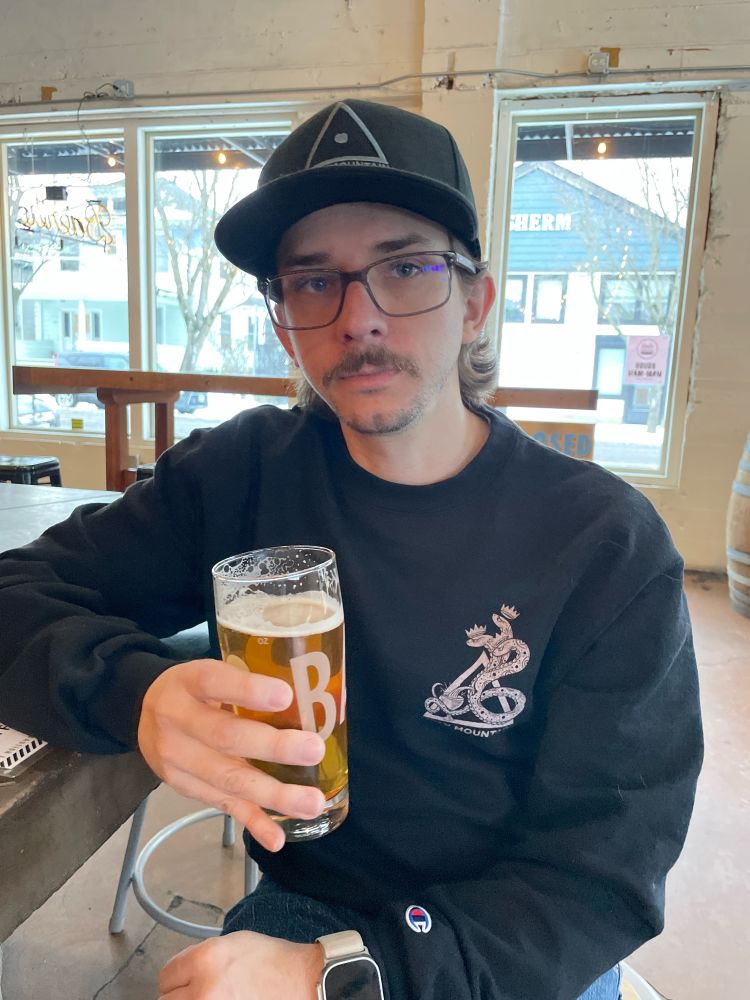 White man holding a beer in a brewery 
wearing a black hat and a black crew neck