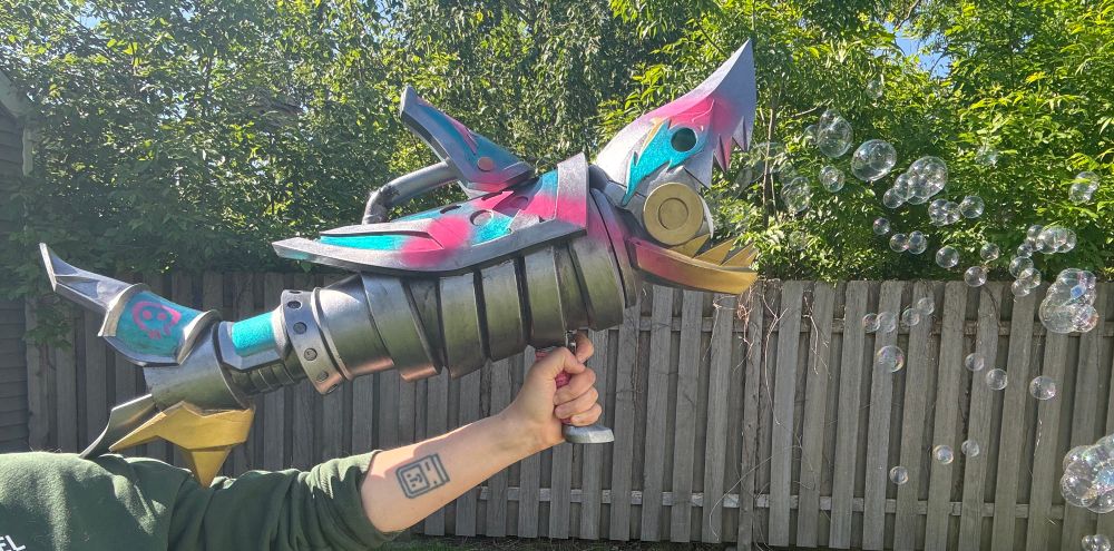 A arm holds a prop- a large, cartoony looking large gun with a shark head and fins shooting bubbles out of its mouth. It is part of a costume for Jinx, from the tv show arcane 