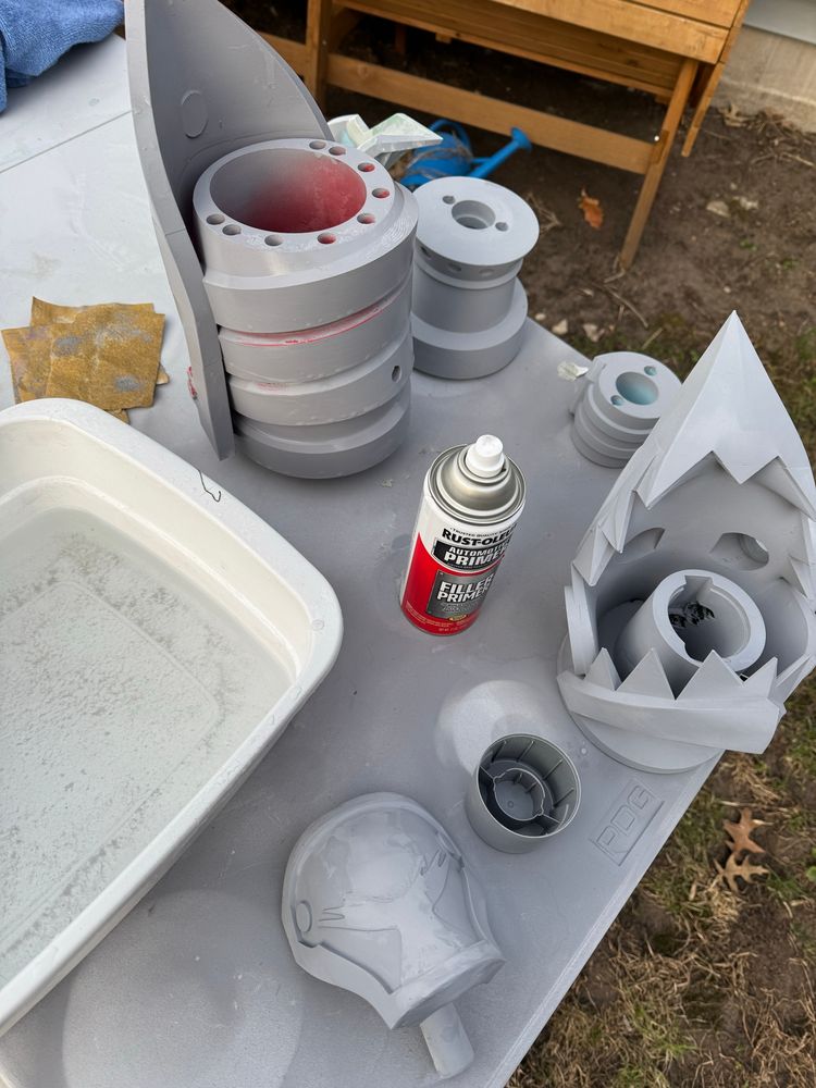 A bunch of parts from the gun fishbones from the tv show arcane sit on a table, painted grey. A can of rust oleum filler-primer is on the table, along with some wet sandpaper and a plastic basin for wet sanding. 