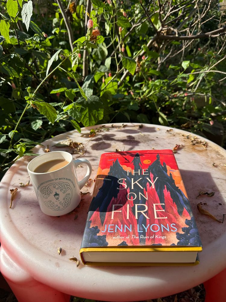 A faded red table with a white mug of tea with green letters and a lantern, next to the book The sky on fire, by Jenn Lyons. The table has some dried leaves scattered around. The sun is casting shadows from the overhead trees.