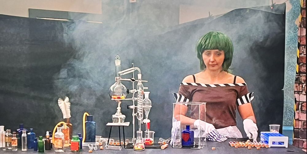 Scottish Willy Wonka Meme. Disinterested actor dressed as an Oompaloompa stands in front of a work table with various laboratory items setup. Entire shot is in front of a flimsy back drop and smoke lingers.