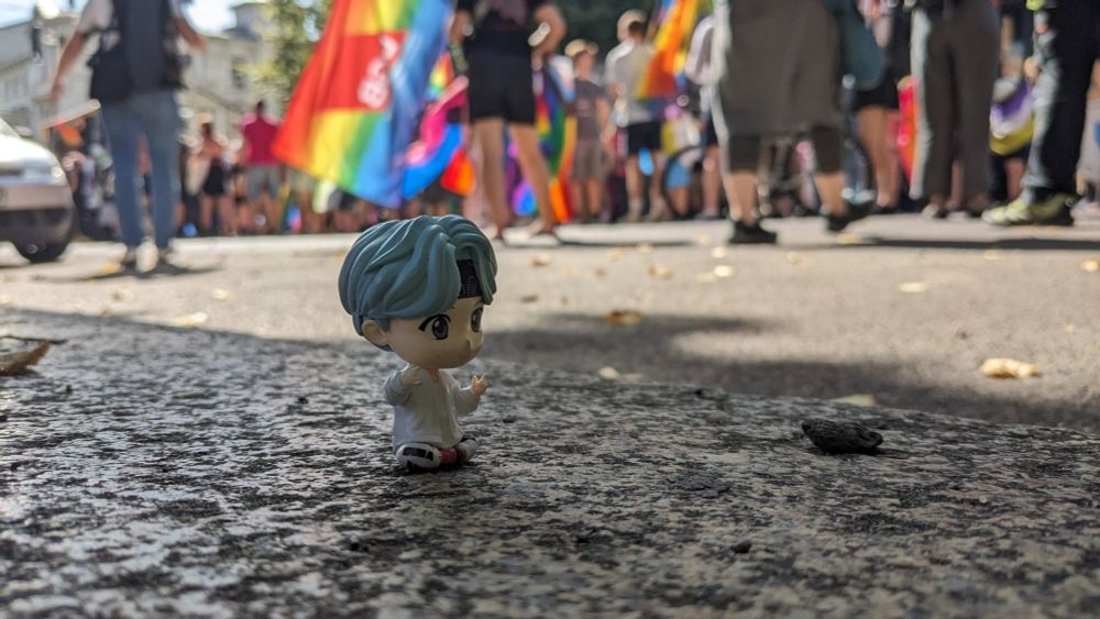 A tiny figure of a person in white clothes and blue hair, depicting BTS member Min Yoongi, sitting on the ground. In the background, there is a crowd of people with some rainbow-colored flags.