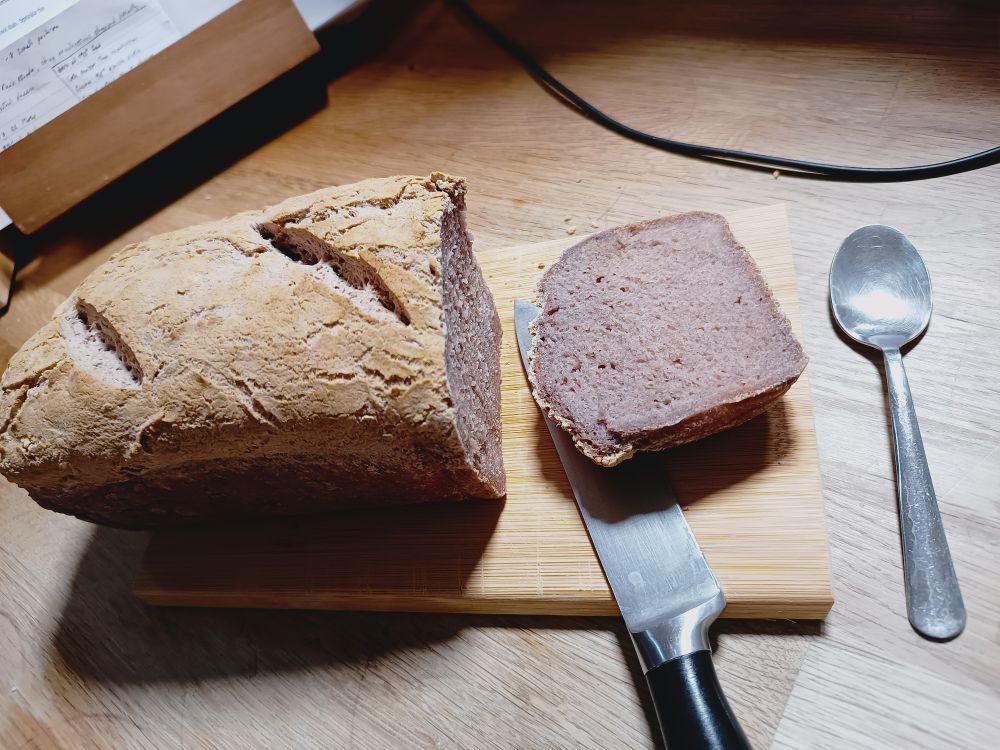 A small loaf of GF bread on a small cutting board, teaspoon for scale. Yes it's small. The inside of the bread shows a pinky-purple colour. Fun.
