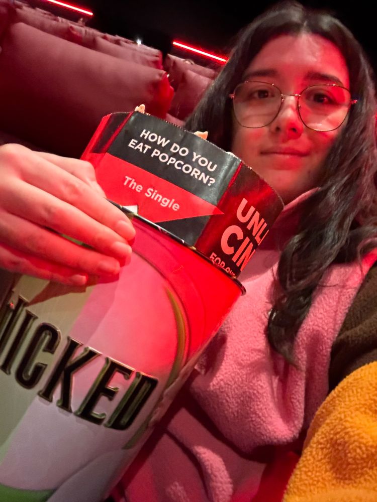 Emma is sitting in the cinema holding her Wicked themed popcorn bucket which is green on one side and pink on the other, and has the Wicked logo across the middle. Inside the bucked is the paper bag of popcorn.