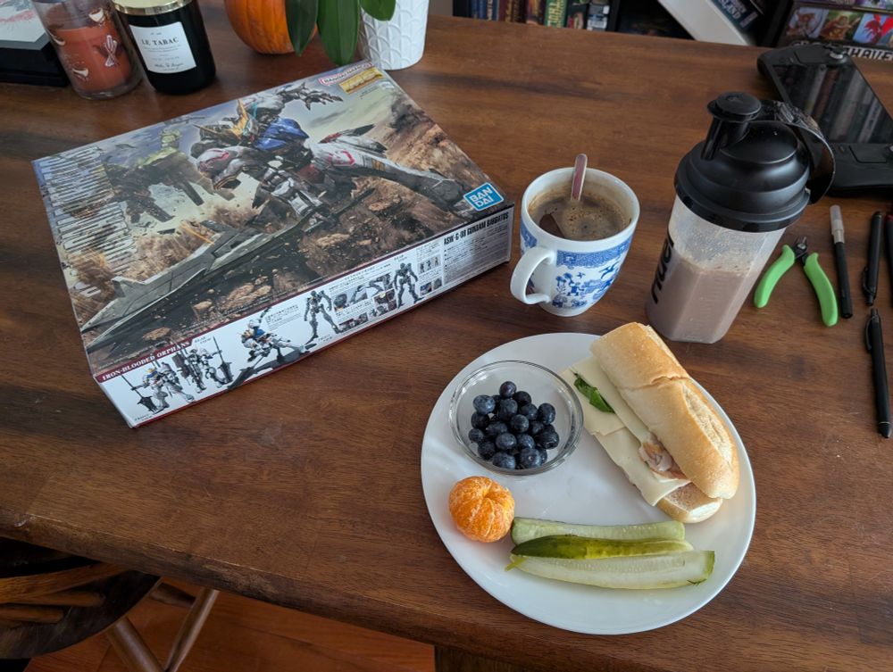 Barbaros Gundam next to a coffee, protein shake, a small bowl of blueberries, pickle slices, mandarin, and turkey sandwich 