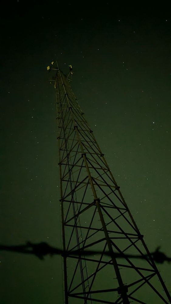 A photo of a radio tower at night. In the foreground a small section of barbed wire can be seen. The picture is angled up in a way that you can see almost the whole tower from top to bottom. The sky has a slight green tint to it 