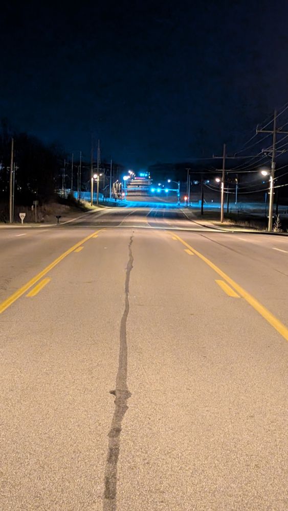 A photo looking down a really long road at night. The entire road is lit up by streetlights and stoplights, but there are no cars to be seen. It is a very liminal photo because it is a 5 lane road, which should normally be filled with traffic.