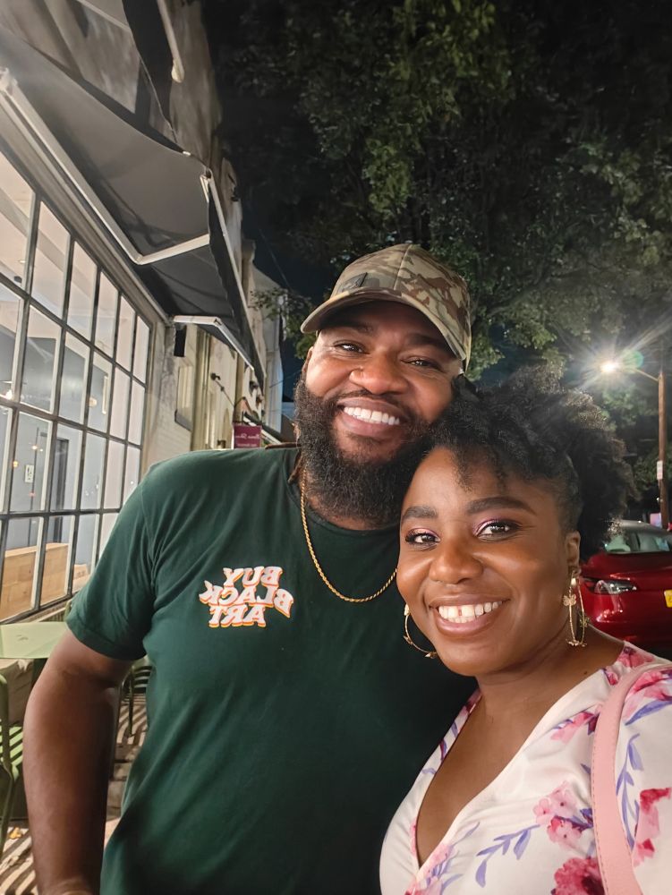 A beautiful, tiny Ghanaian woman poses with a gnarly, self-proclaimed tall South Carolinian man. She adorns a floral dress and he just want yall niggas to but Black art. 