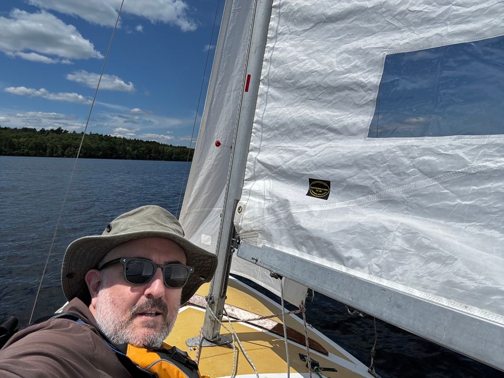 A man wearing sunglasses and a hat on a small sailboat on a lake with blue skies