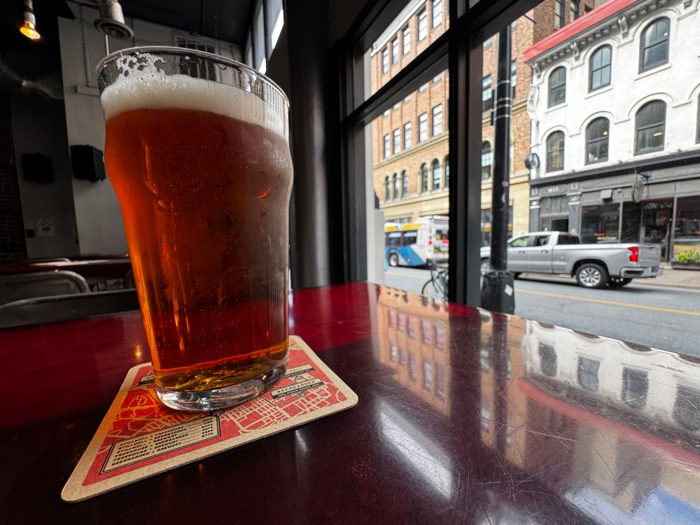 A copper-coloured pint glass of beer rests on a dark red table with a busy street outside