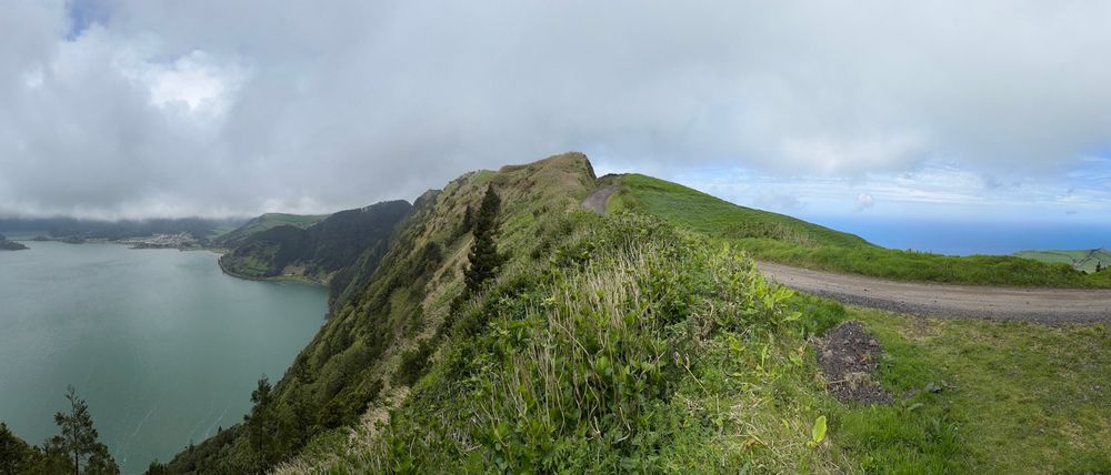 A panoramic photo taken from a high vantage point on the rim of a volcano, a hiking trail is seen in the middle, the crater lake is seen in the left, the Atlantic Ocean on the right