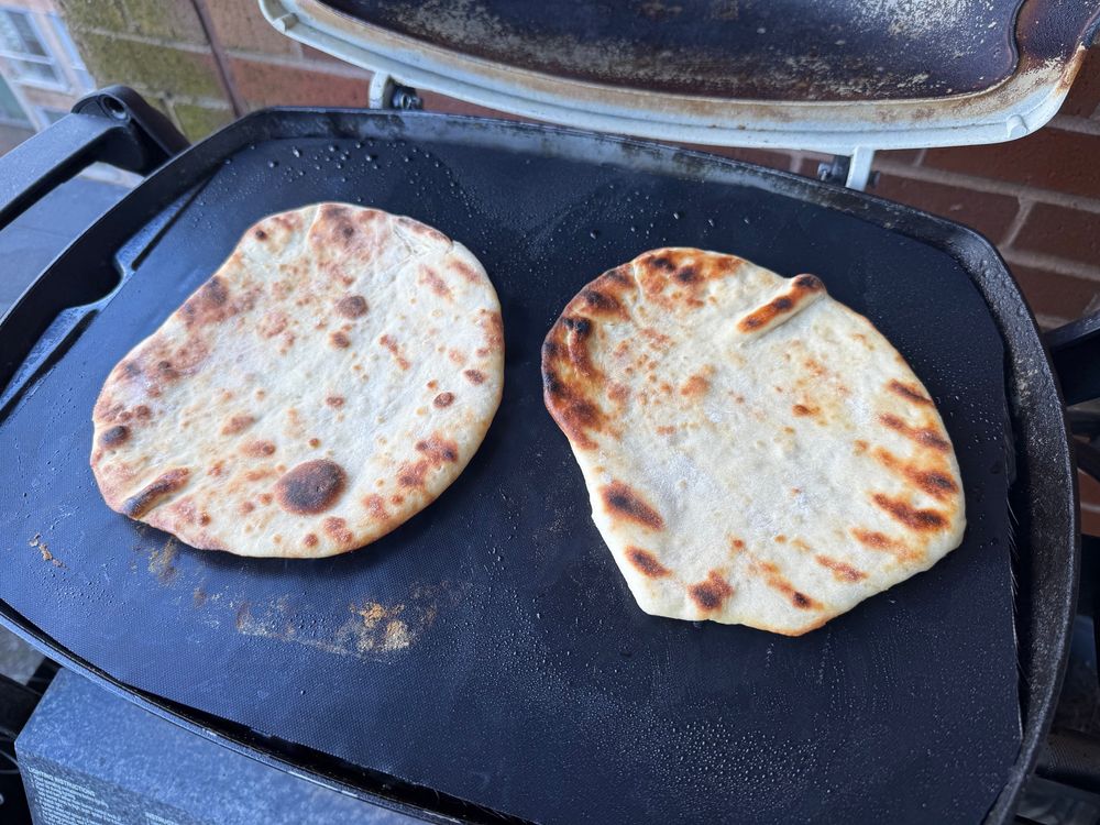 Two roughly circular flat breads are baking on a bbq and have turned a perfect shade of brown