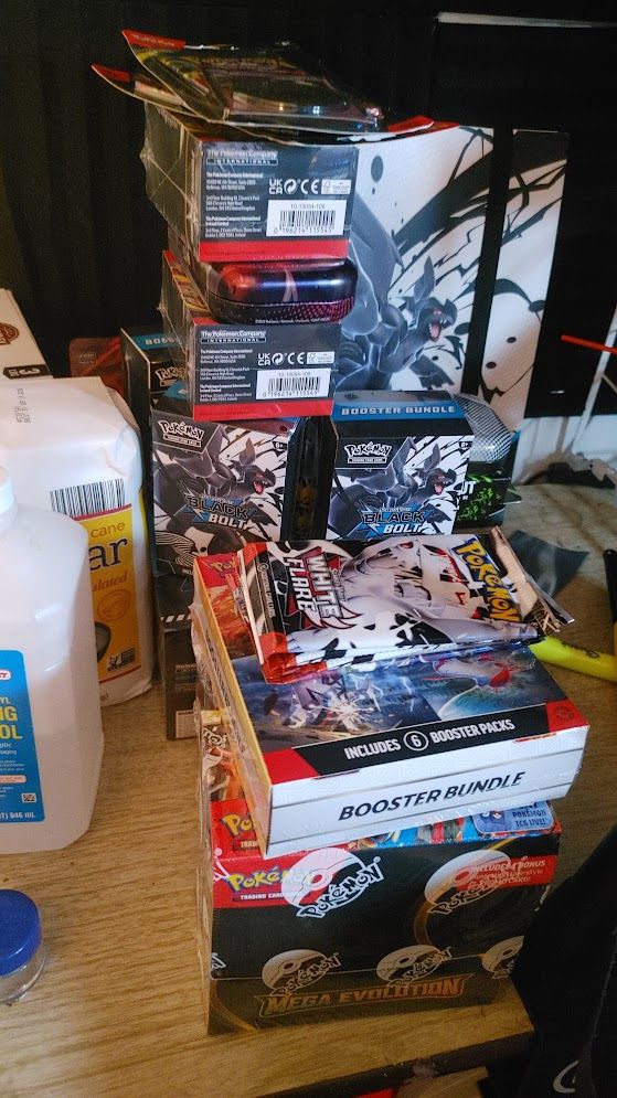 A pile of Pokémon TCG boxes, including Surging Sparks, Mega Evolution, Black Bolt, and some loose packs! Also there's a 1lb bag of sugar, and some rubbing alcohol off to the side. Don't worry about that *smile*