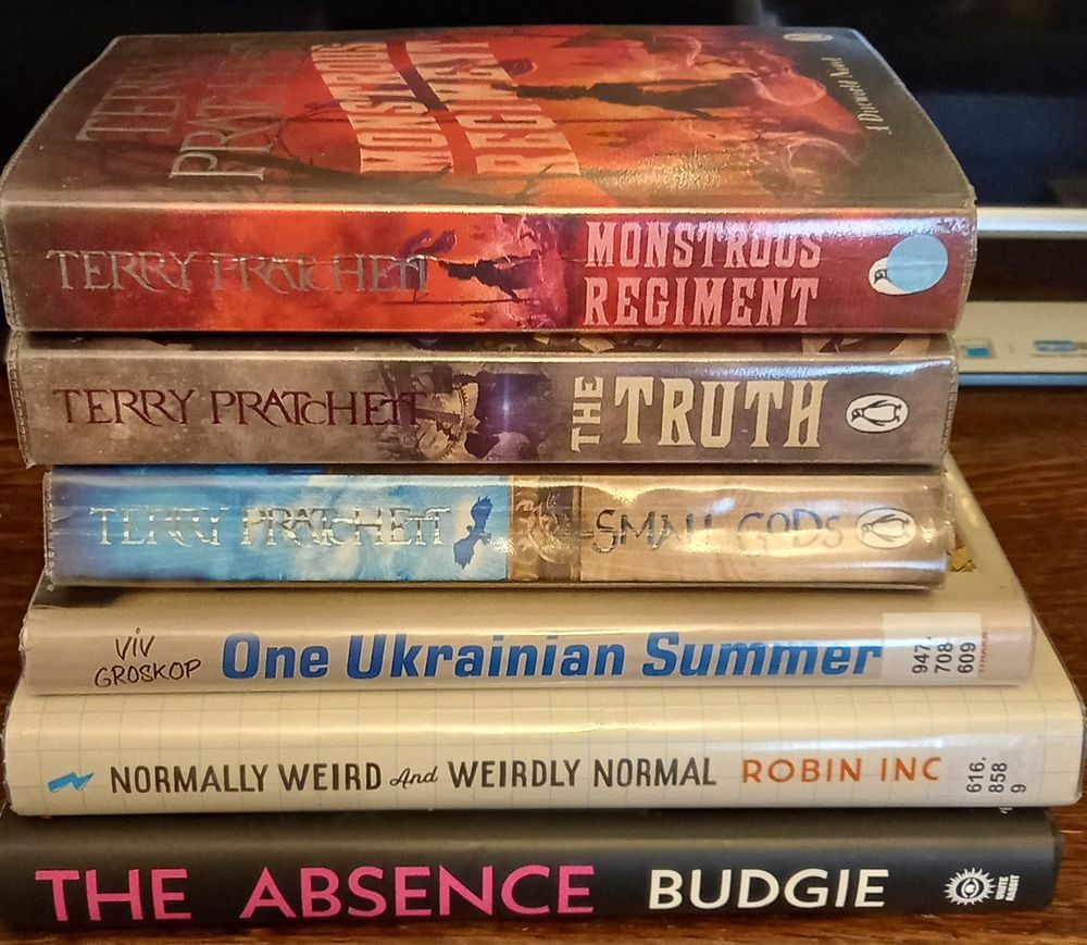 A pile of books: Normally Weird and Weirdly Normal by Robin Ince, One Ukrainian Summer by  Viv Groskop, The Absence by Budgie, The Truth by Terry Pratchett, Monstrous Regiment by Terry Pratchett, Small Gods by Terry Pratchett.