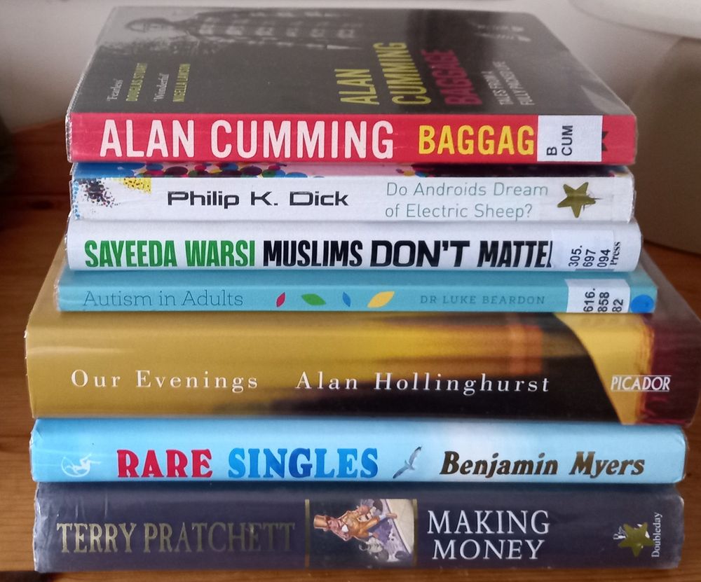 A pile of library books: Baggage by Alan Cumming, Do Androids Dream of Electric Sheep by Philip K Dick, Muslims Don’t Matter by Sayeeda Warsi, Autism in Adults by Luke Beardon, Our Evenings by Alan Hollinghurst, Rare Singles by Benjamin Myers, Making Money by Terry Pratchett.