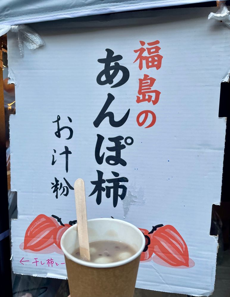 紙カップに入ったお汁粉の写真です。お汁粉の中に白玉や小豆やカットされたあんぽ柿が入っています。学祭の屋台の看板が奥にみえます。