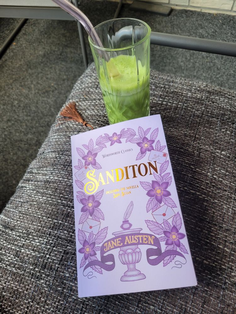 Ein iced matcha und ein Buch auf einem grau melliertem Kissen. Das Buch ist "Sanditon including the novella Lady Susan" (ein Buch mit nicht veröffentlichten Kurzgeschichten und unvollendeten Romanen) von Jane Austen. 