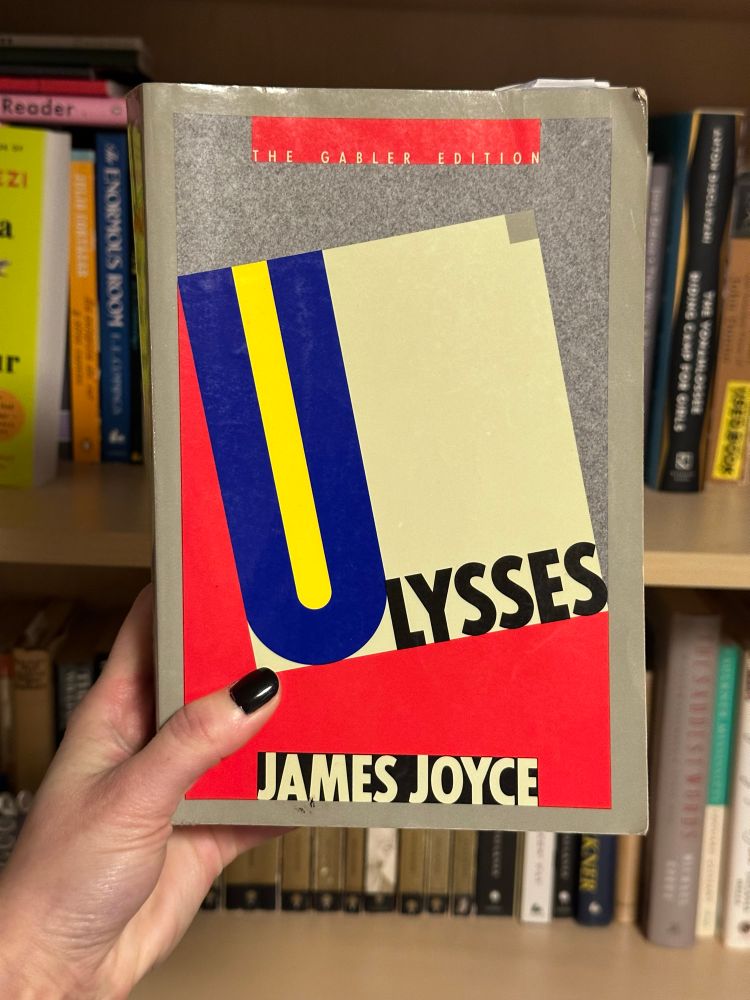 Hand holding a copy of Ulysses by James Joyce in front of a bookshelf. The cover artwork has a red, yellow, and blue color block design.